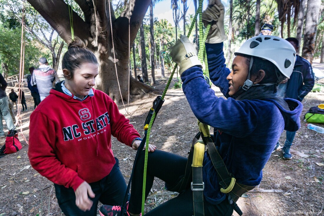 A woman stands in front of another woman who is in a climbing harness, teaching her how to climb a tree. 